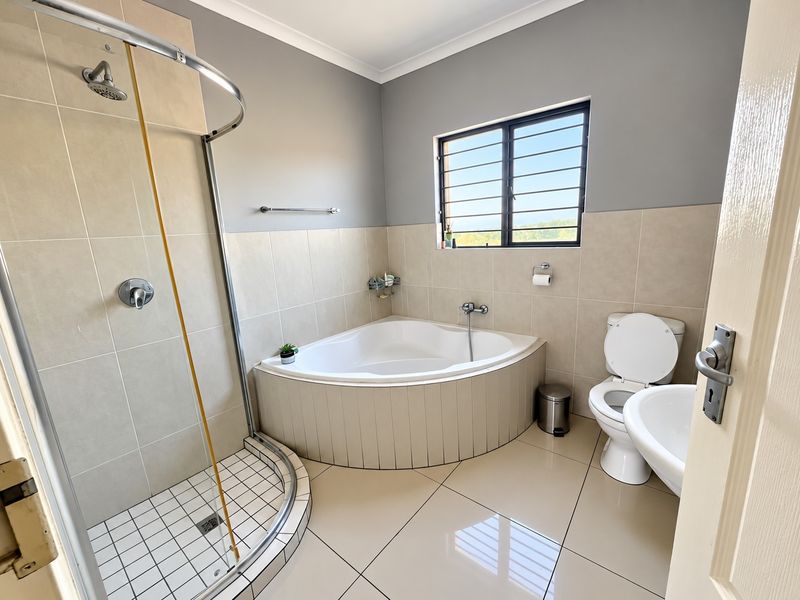 A clean, well-maintained bathroom featuring a corner bathtub, separate shower enclosure, pedestal basin, and neutral tiled finishes with security bars on the window.