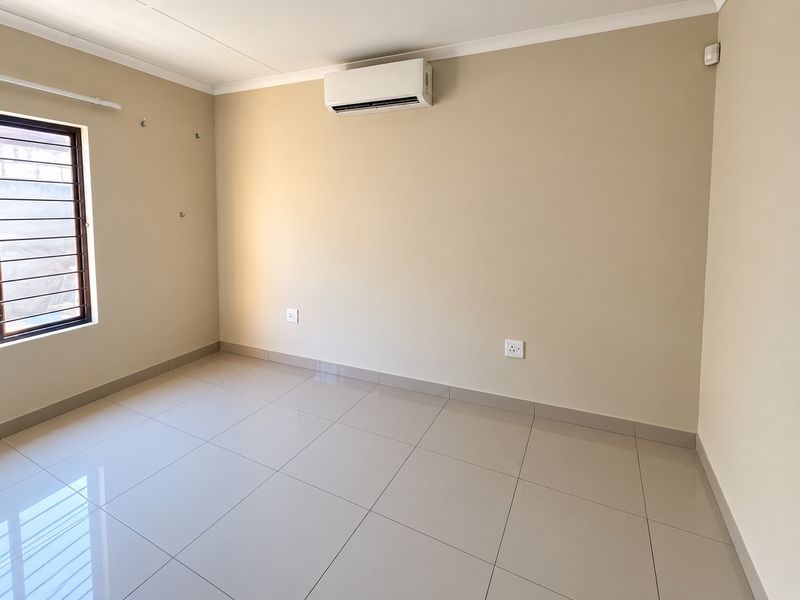 A clean, empty bedroom featuring neutral tiled flooring, a wall-mounted air conditioning unit, and security bars on the window.