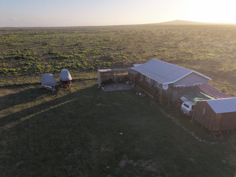 Aerial view of a rural property featuring a main dwelling with a corrugated metal roof, attached patio, and surrounding open land.
