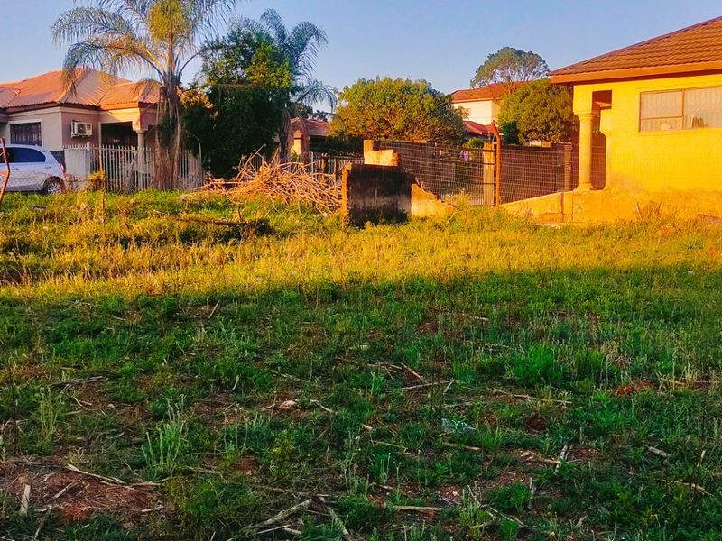 A vacant residential plot featuring overgrown grass, damaged wire fencing, and a partial concrete boundary wall in a suburban neighborhood setting.