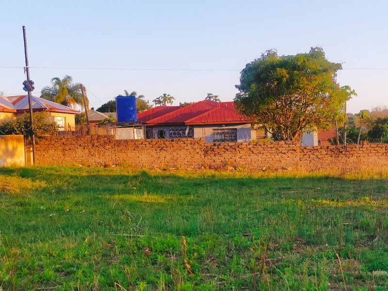A view of a vacant residential plot featuring a brick boundary wall with security fencing and neighboring properties in the background.
