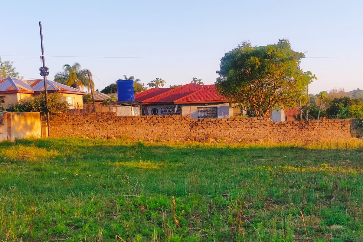 A view of a vacant residential plot featuring a brick boundary wall with security fencing and neighboring properties in the background.