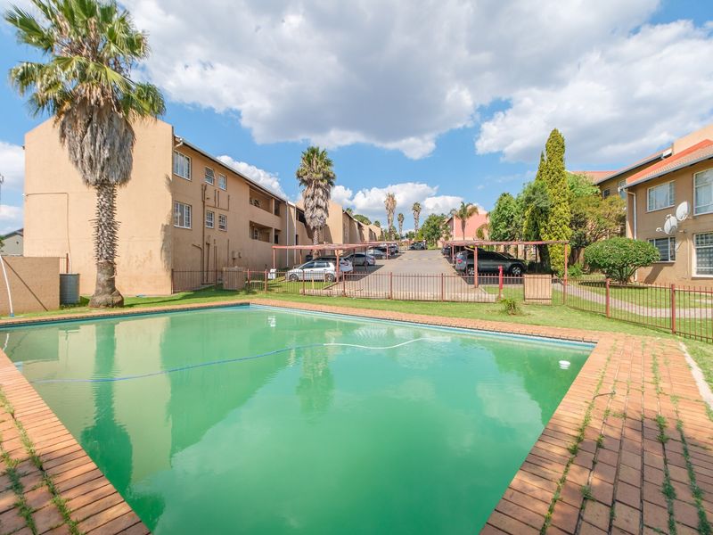 Communal swimming pool area featuring brick paving and metal fencing, situated within an apartment complex with covered parking bays.