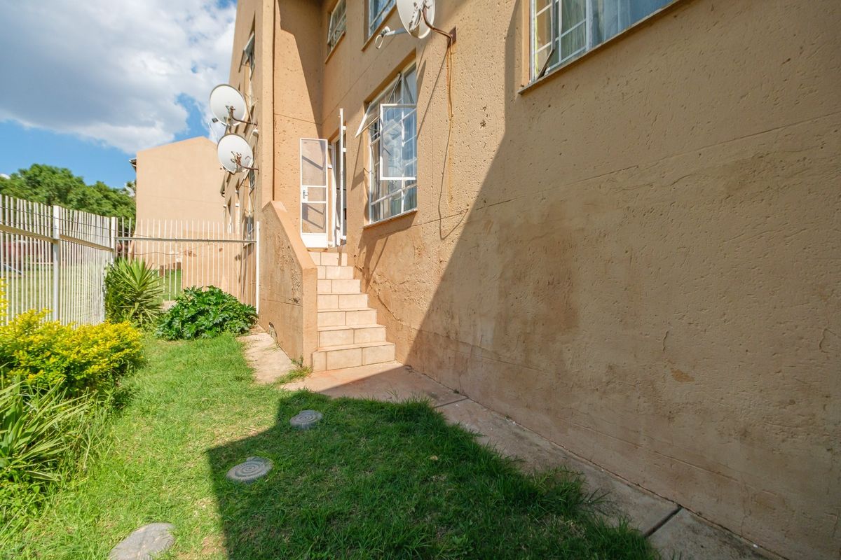 Rear exterior view of the apartment building featuring a tiled staircase entrance, security gate, and surrounding garden area with metal fencing.