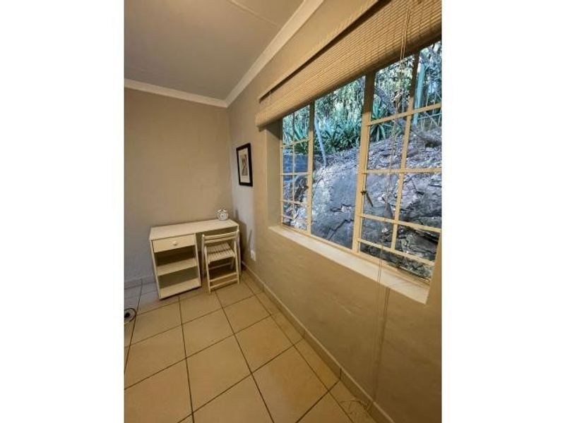 A functional bedroom featuring tiled flooring, a large window with a bamboo blind, and neutral wall finishes, offering a view of the natural rock embankment.