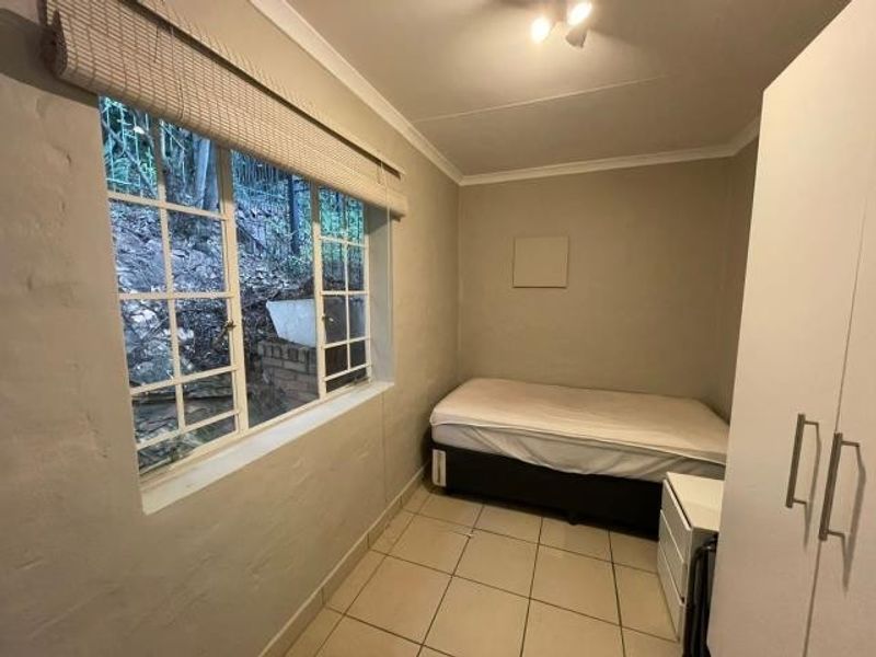 A compact bedroom featuring tiled flooring, a built-in wardrobe, and a window with security bars, offering a functional and neutral living space.
