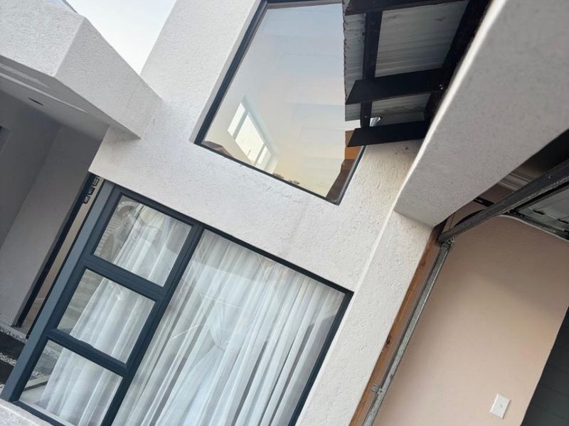 Modern double-storey residential facade featuring large aluminium-framed windows, textured plaster finish, and a paved driveway entrance.