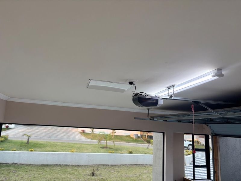 A clean, well-lit garage interior featuring a mounted garage door motor, ceiling lighting, and a large window overlooking the front yard.