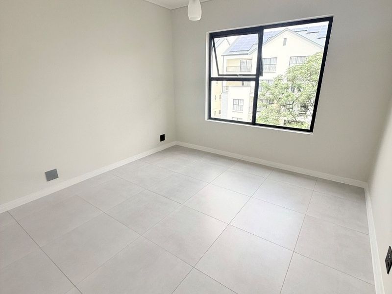 A bright, empty bedroom featuring neutral tiled flooring, clean white walls, and a modern black-framed window with natural light.