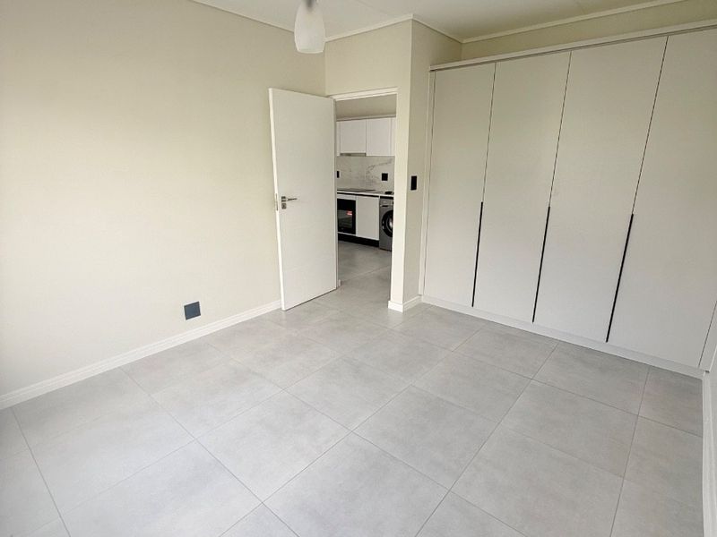 A bright, empty bedroom featuring modern light-grey floor tiles, ample built-in cupboard space, and a clean, neutral aesthetic with direct access to the kitchen area.
