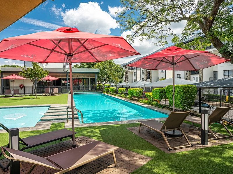A well-maintained communal swimming pool area featuring paved decking, artificial grass, and established landscaping within an apartment complex.