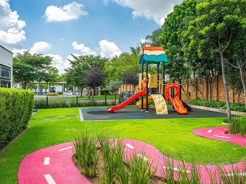 A well-maintained communal garden area featuring a children's playground with rubberized flooring, landscaped lawns, and a decorative paved pathway.