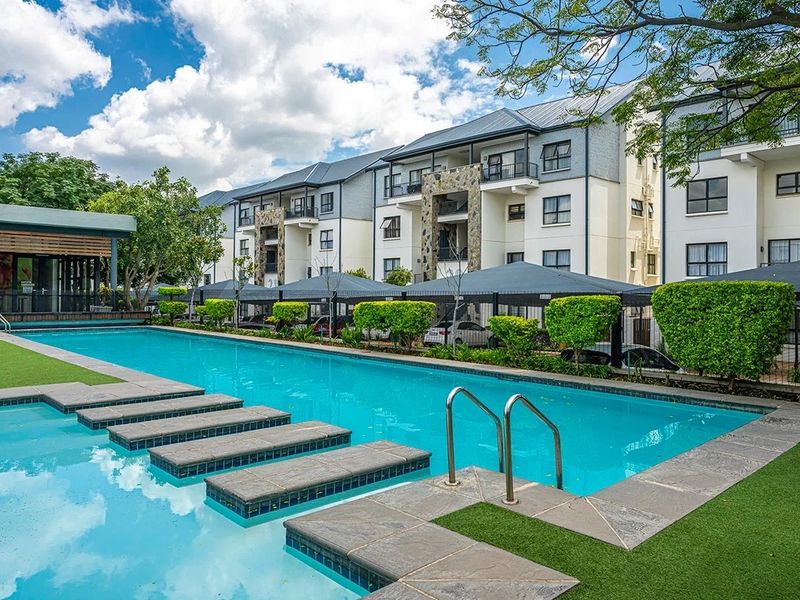 A well-maintained communal swimming pool area featuring stone-clad building facades, landscaped greenery, and covered parking bays within the apartment complex.