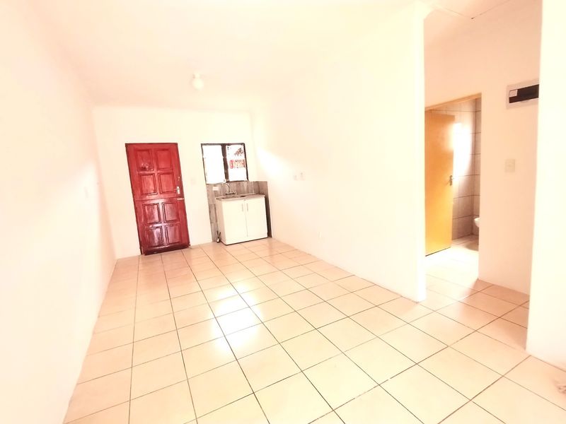 An open-plan living area featuring tiled flooring, a compact kitchen sink unit, and access to an adjoining room.