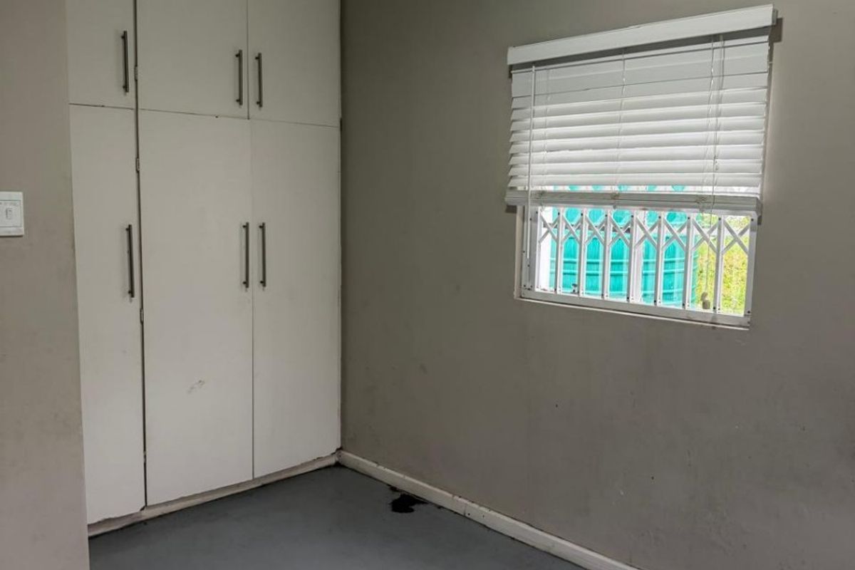 An empty bedroom featuring built-in cupboards, a window with venetian blinds and security gate, and painted concrete flooring.