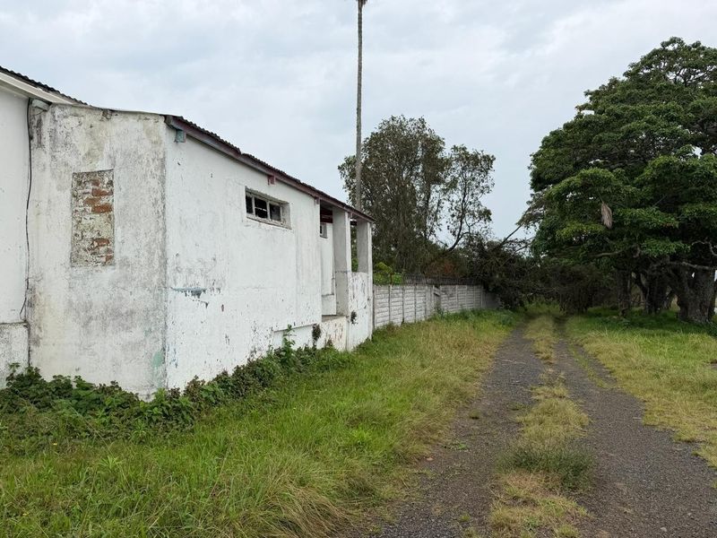 Exterior view of a small holding structure showing significant wear to the exterior walls, a gravel driveway, and surrounding overgrown garden area.