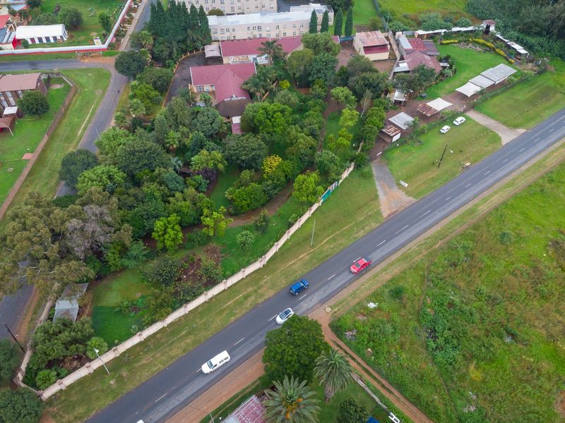 Aerial view of a large farm property featuring extensive mature gardens, multiple roof structures, and a perimeter wall adjacent to a public road.
