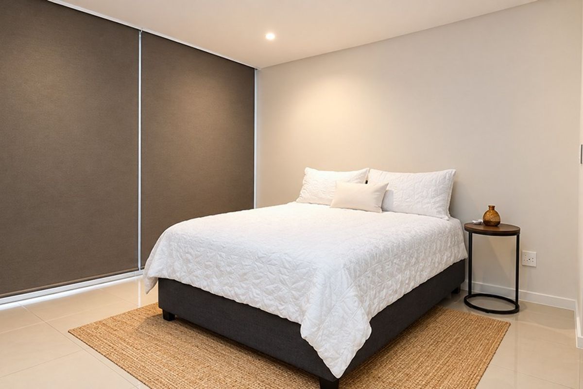 A modern, well-maintained bedroom featuring neutral tiled flooring, recessed lighting, and large roller blinds for privacy.