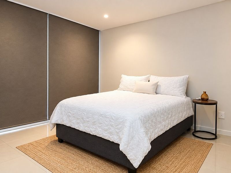 A modern, well-maintained bedroom featuring neutral tiled flooring, recessed lighting, and large roller blinds for privacy.