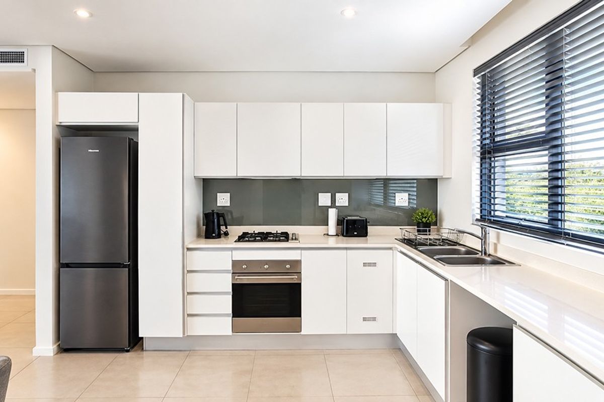Modern kitchen featuring sleek white cabinetry, stone countertops, a gas hob, built-in oven, and a glass splashback, complemented by recessed lighting and large windows.