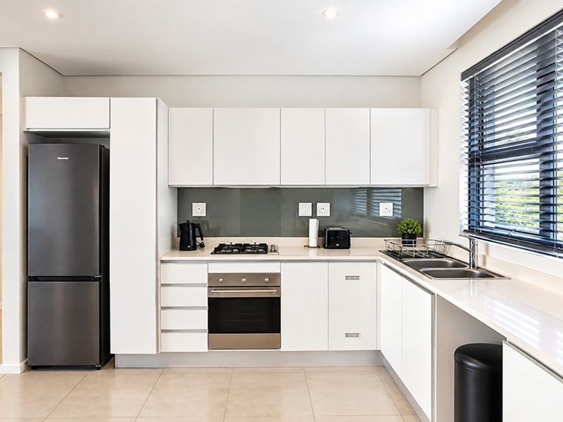 Modern kitchen featuring sleek white cabinetry, stone countertops, a gas hob, built-in oven, and a glass splashback, complemented by recessed lighting and large windows.