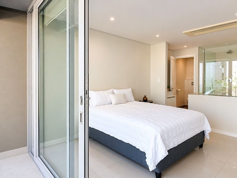 Modern bedroom featuring tiled flooring, recessed lighting, ducted air conditioning, and an open-plan en-suite bathroom with a glass partition, leading to a private balcony.