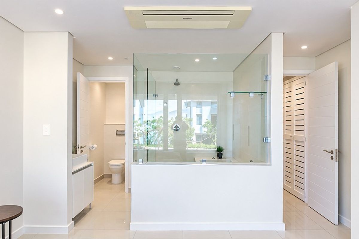 Modern, bright bathroom featuring a glass-enclosed shower, wall-mounted toilet, vanity unit, and integrated ceiling air conditioning system with clean, neutral finishes.
