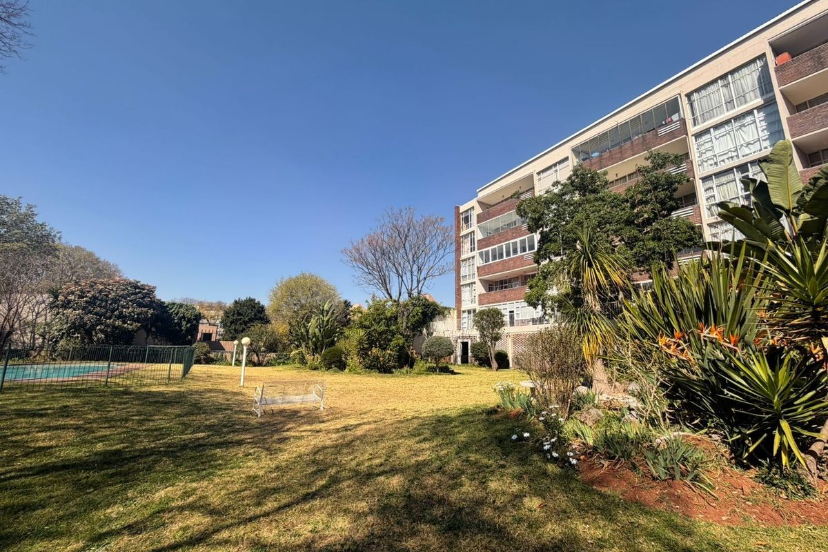 A view of the apartment building exterior featuring a well-maintained communal garden area and a fenced swimming pool.