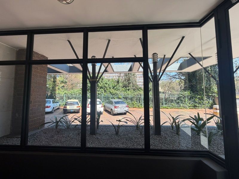A view from inside looking out through large black-framed windows towards a landscaped parking area with structural pillars.