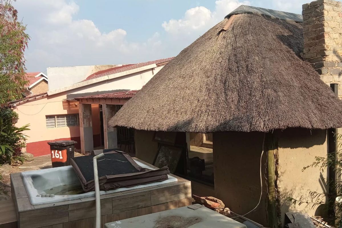 Rear exterior view featuring a thatched lapa structure, a tiled spa, and a brick chimney, showing significant signs of wear and maintenance requirements.