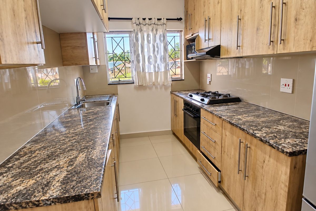 Modern galley-style kitchen featuring wood-look cabinetry, granite countertops, built-in oven, gas hob, and extractor fan with tiled flooring and security bars on windows.