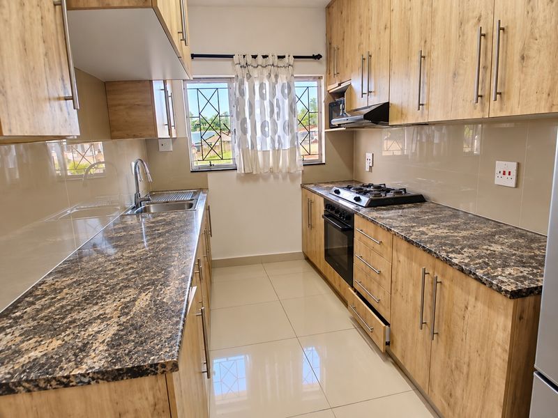 Modern galley-style kitchen featuring wood-look cabinetry, granite countertops, built-in oven, gas hob, and extractor fan with tiled flooring and security bars on windows.