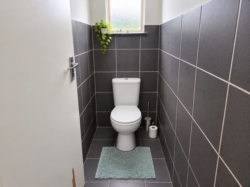 A compact bathroom featuring dark grey wall and floor tiles, a white toilet, and an obscured glass window for privacy.