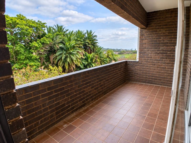 A spacious covered balcony featuring exposed brick walls and tiled flooring, offering an elevated view of the surrounding greenery.