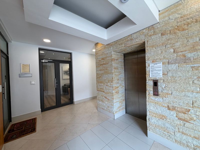 Modern office building lobby featuring a stainless steel elevator, natural stone feature wall, recessed ceiling lighting, and tiled flooring.