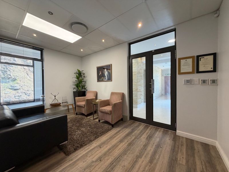 A professional office reception area featuring modern laminate flooring, a suspended ceiling with recessed lighting, and a secure double glass entrance door.