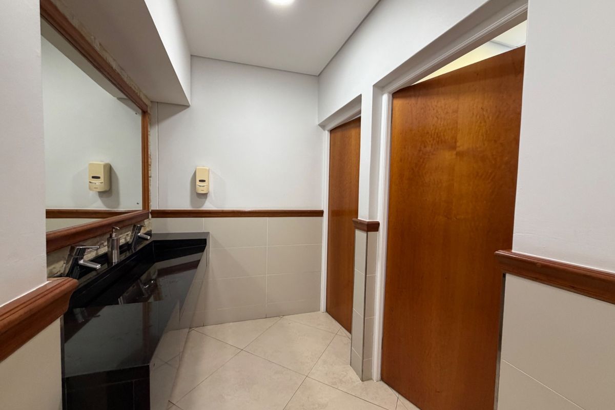 A clean and functional office bathroom featuring a black granite vanity, large mirror, and neutral ceramic tiling with wooden door finishes.
