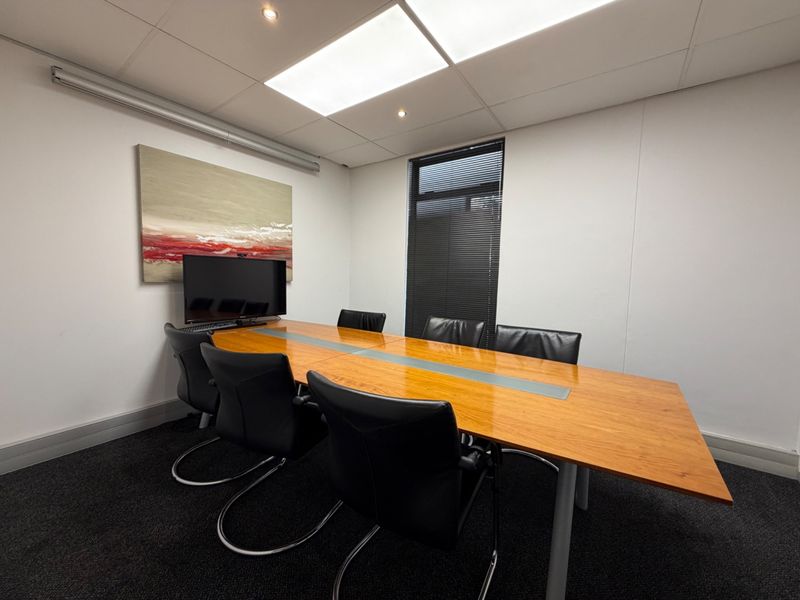 A professional office meeting room featuring a large boardroom table, recessed ceiling lighting, and a window with fitted blinds.