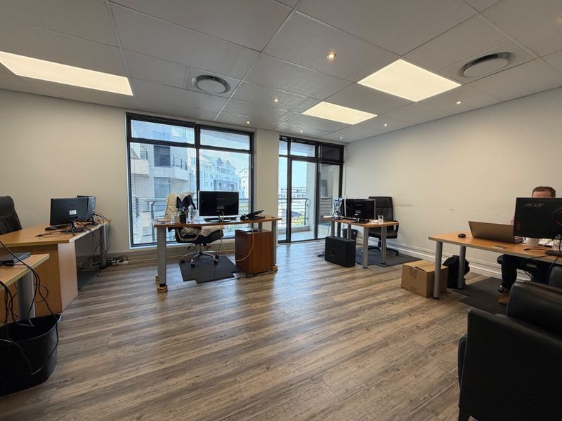 A functional open-plan office space featuring laminate flooring, suspended ceilings with recessed lighting, and large windows providing natural light and balcony access.