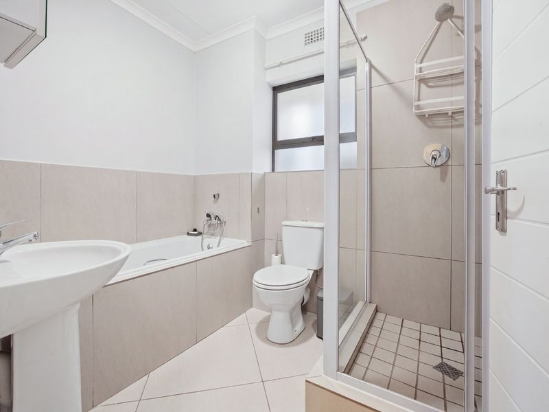 A clean, modern bathroom featuring a pedestal basin, a built-in bathtub, and a separate glass-enclosed shower with neutral wall and floor tiling.