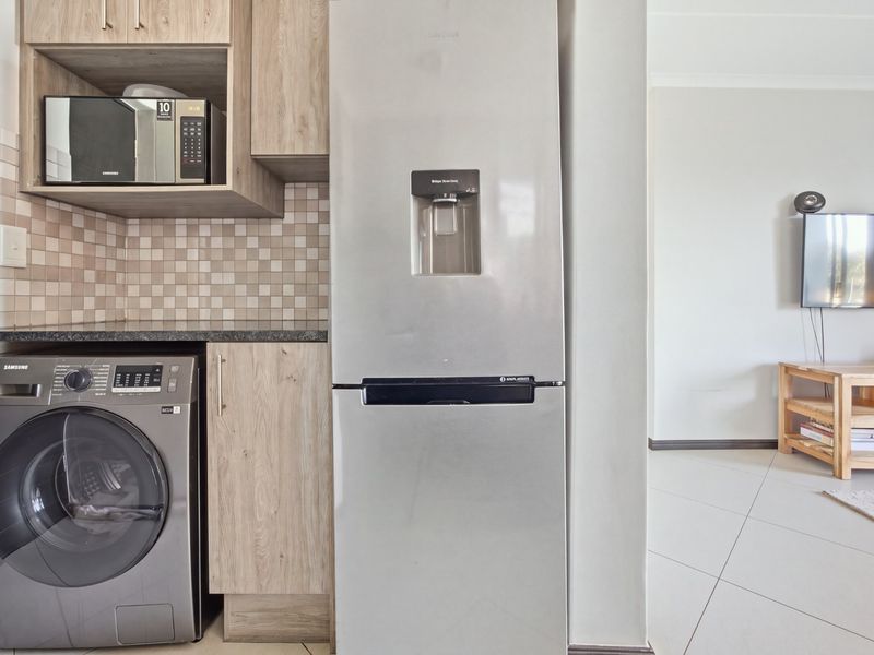 A modern kitchen area featuring light wood-toned cabinetry, a tiled backsplash, and integrated laundry appliances, seamlessly flowing into the adjacent living space.