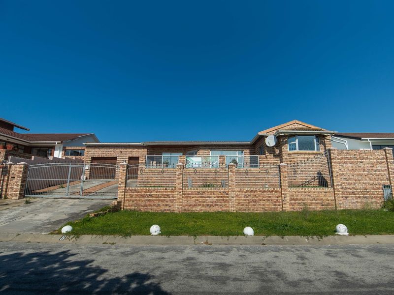 Front elevation of a face-brick house featuring a perimeter wall, metal driveway gate, and paved access, set against a clear blue sky.