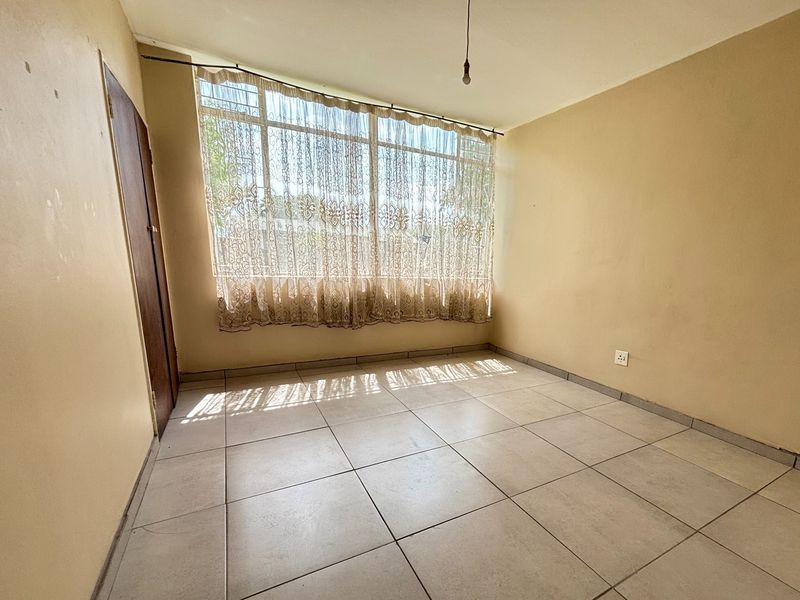 An empty bedroom featuring neutral tiled flooring, a large window allowing natural light, and plain walls ready for personalization.