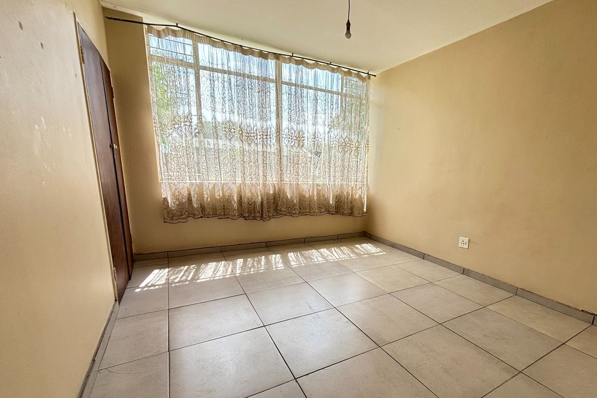 An empty bedroom featuring neutral tiled flooring, a large window allowing natural light, and plain walls ready for personalization.