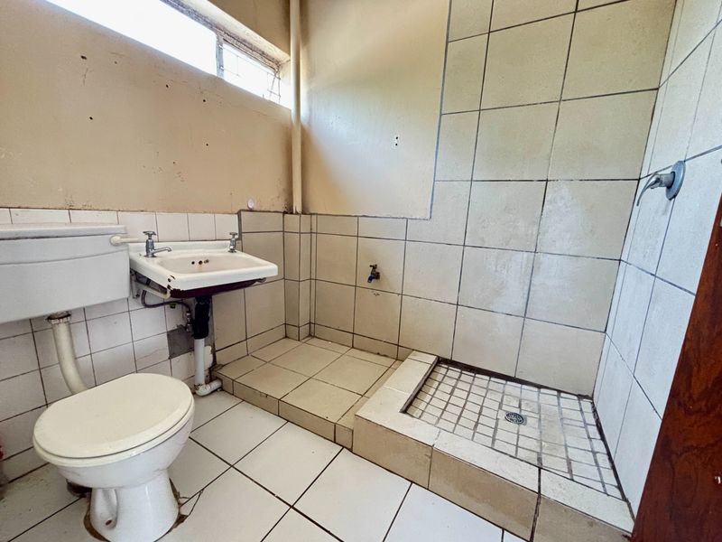 A dated bathroom featuring a toilet, wall-mounted basin, and a tiled shower area, requiring significant renovation and cosmetic updates to improve functionality and aesthetics.