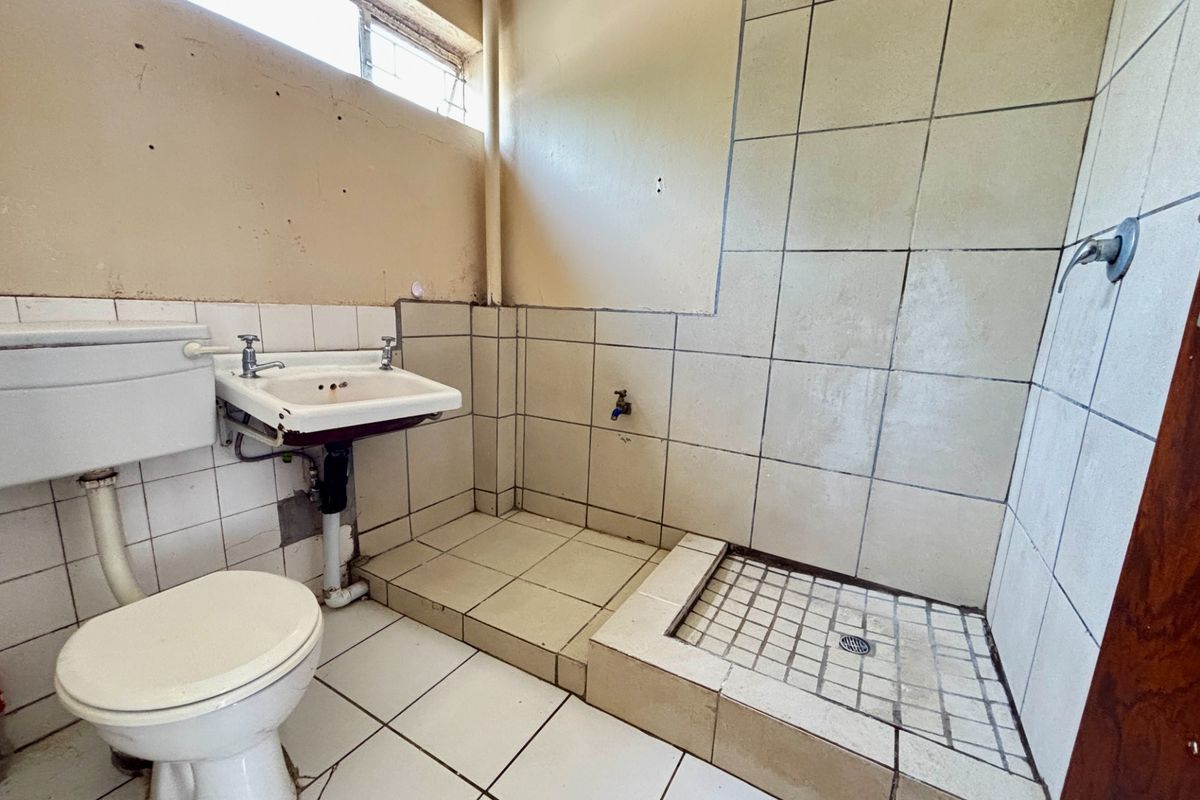 A dated bathroom featuring a toilet, wall-mounted basin, and a tiled shower area, requiring significant renovation and cosmetic updates to improve functionality and aesthetics.