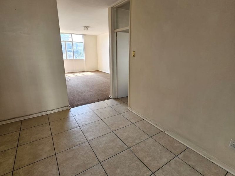 An empty, open-plan living area featuring a combination of tiled and carpeted flooring, neutral wall finishes, and natural light from a large window.
