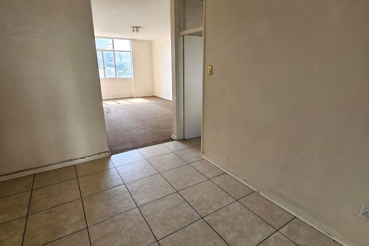 An empty, open-plan living area featuring a combination of tiled and carpeted flooring, neutral wall finishes, and natural light from a large window.