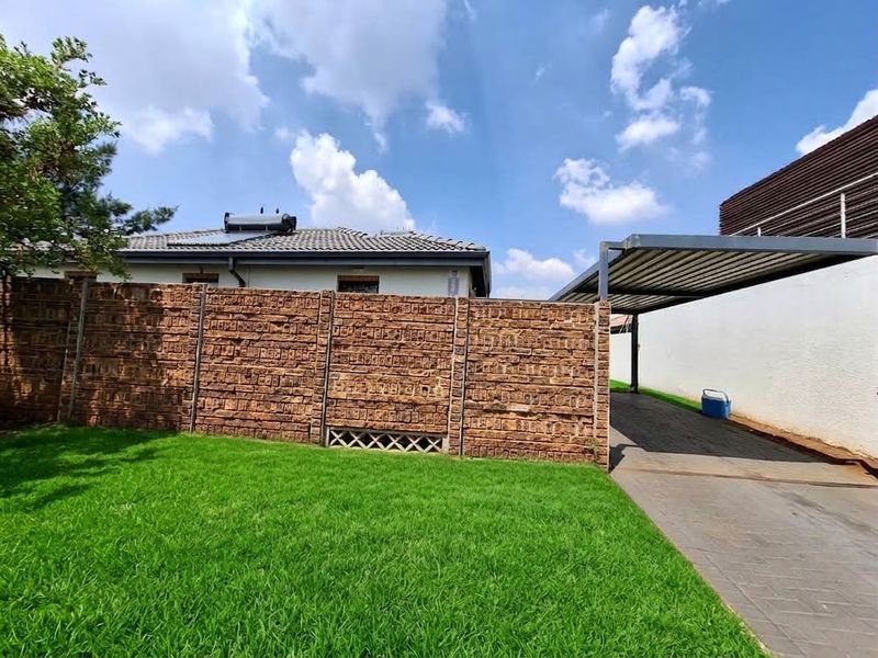 Rear garden area featuring a well-maintained lawn, brick boundary wall, paved driveway, and a covered carport structure.