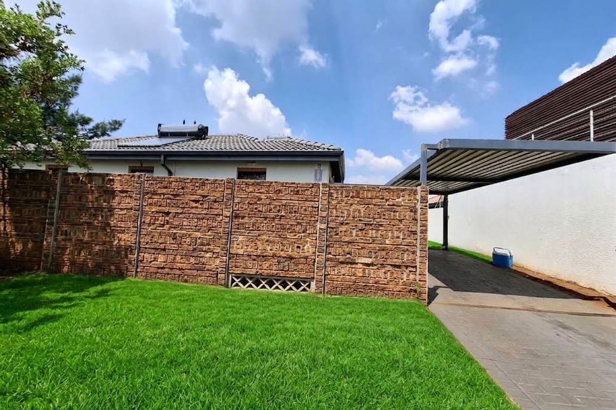 Rear garden area featuring a well-maintained lawn, brick boundary wall, paved driveway, and a covered carport structure.